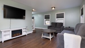 A living room with a fireplace, a television mounted on the wall, a grey sofa, and a round coffee table. - Convenient location for adventures in the Adirondacks! (Warrensburg)