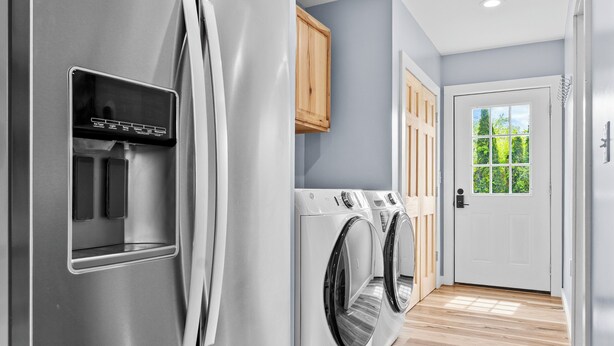 Une cuisine moderne dotée d’appareils électroménagers en acier inoxydable, d’un lave-linge et d’un sèche-linge intégrés, de planchers en bois et offrant une vue sur un espace extérieur verdoyant, accessible par une porte.