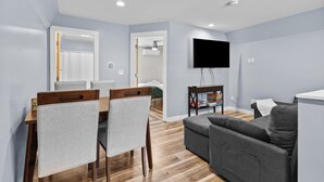 Un salon moderne avec un canapé gris, une table à manger en bois avec des chaises, un téléviseur à écran plat fixé au mur et un ventilateur de plafond en arrière-plan.