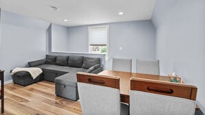 Un salon moderne avec un canapé d’angle, une table à manger en bois et quatre chaises, ainsi qu’une fenêtre équipée de stores.