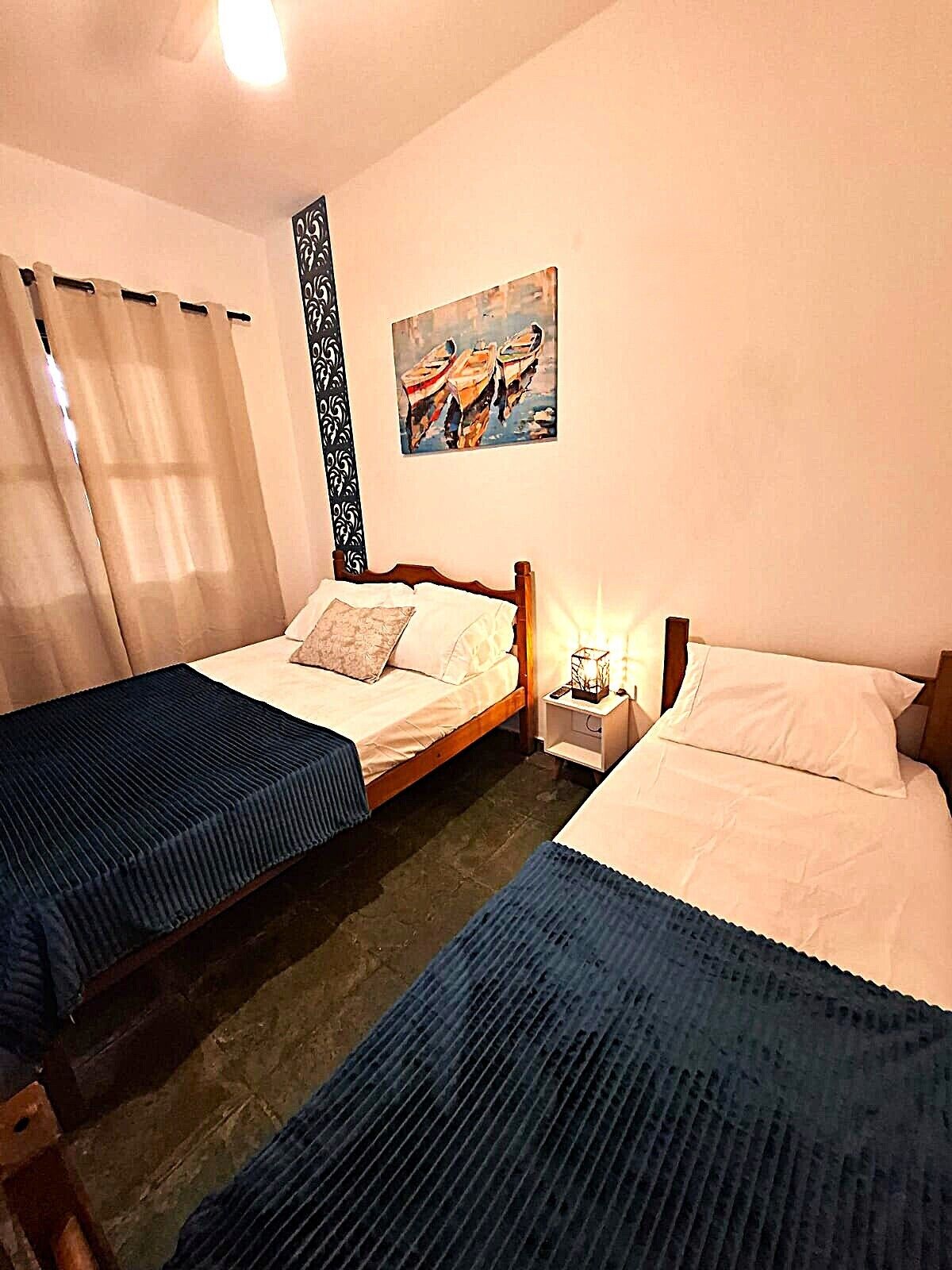 A double room with two beds, a nightstand with a lamp, a painting on the wall, and a window with curtains.
