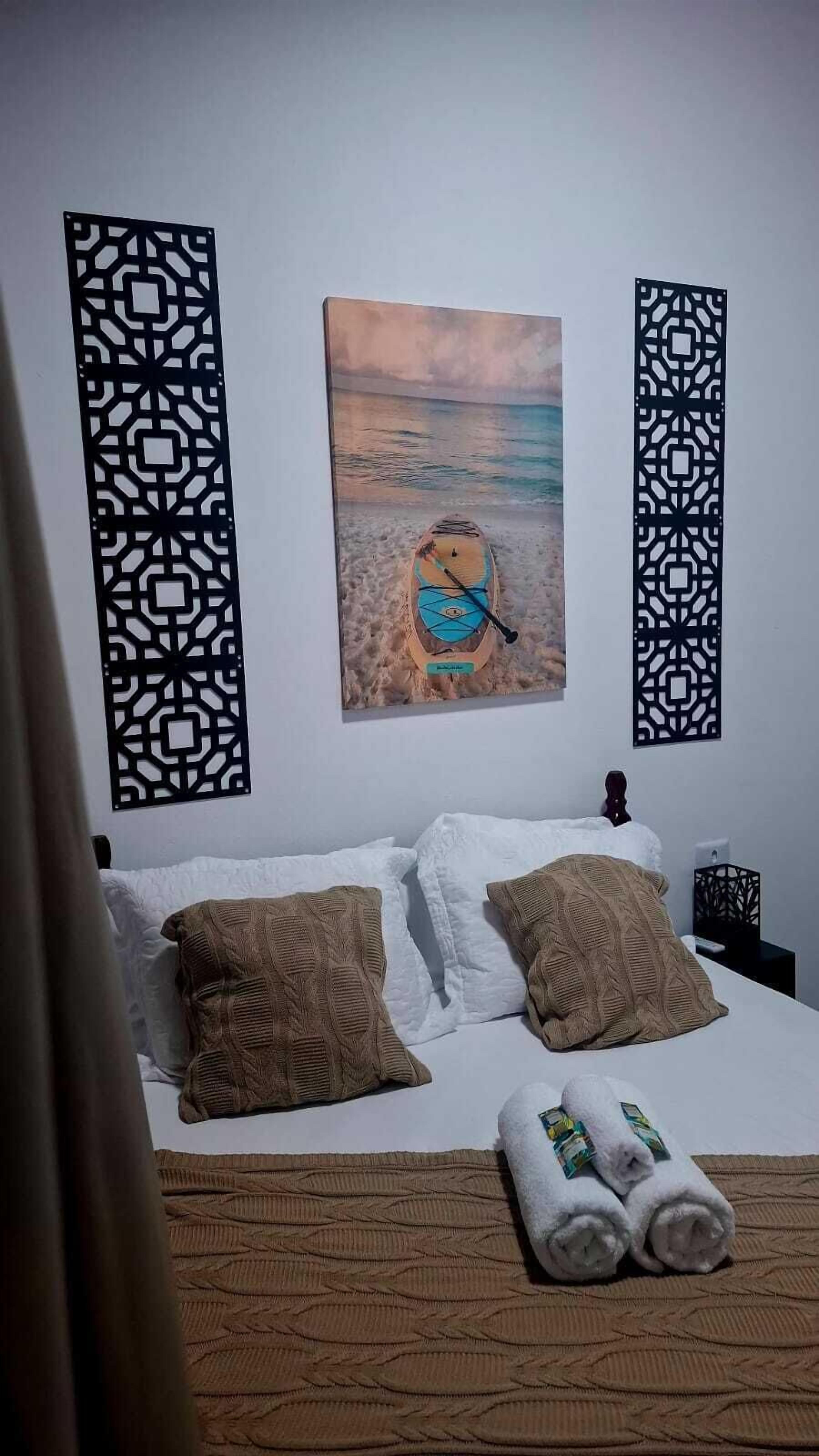 Habitación de hotel con cama, paneles decorativos, un cuadro con escena de playa y toallas sobre la cama.