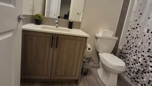 Een moderne badkamer met een houten wastafelmeubel, toilet, douche met een decoratieve gordijn en een spiegel met verlichting erboven.