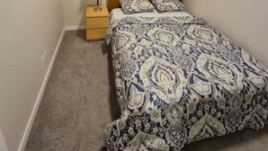 Een slaapkamer met een bed, nachtkastje, lamp, raam met jaloezieën en een lijst aan de muur.