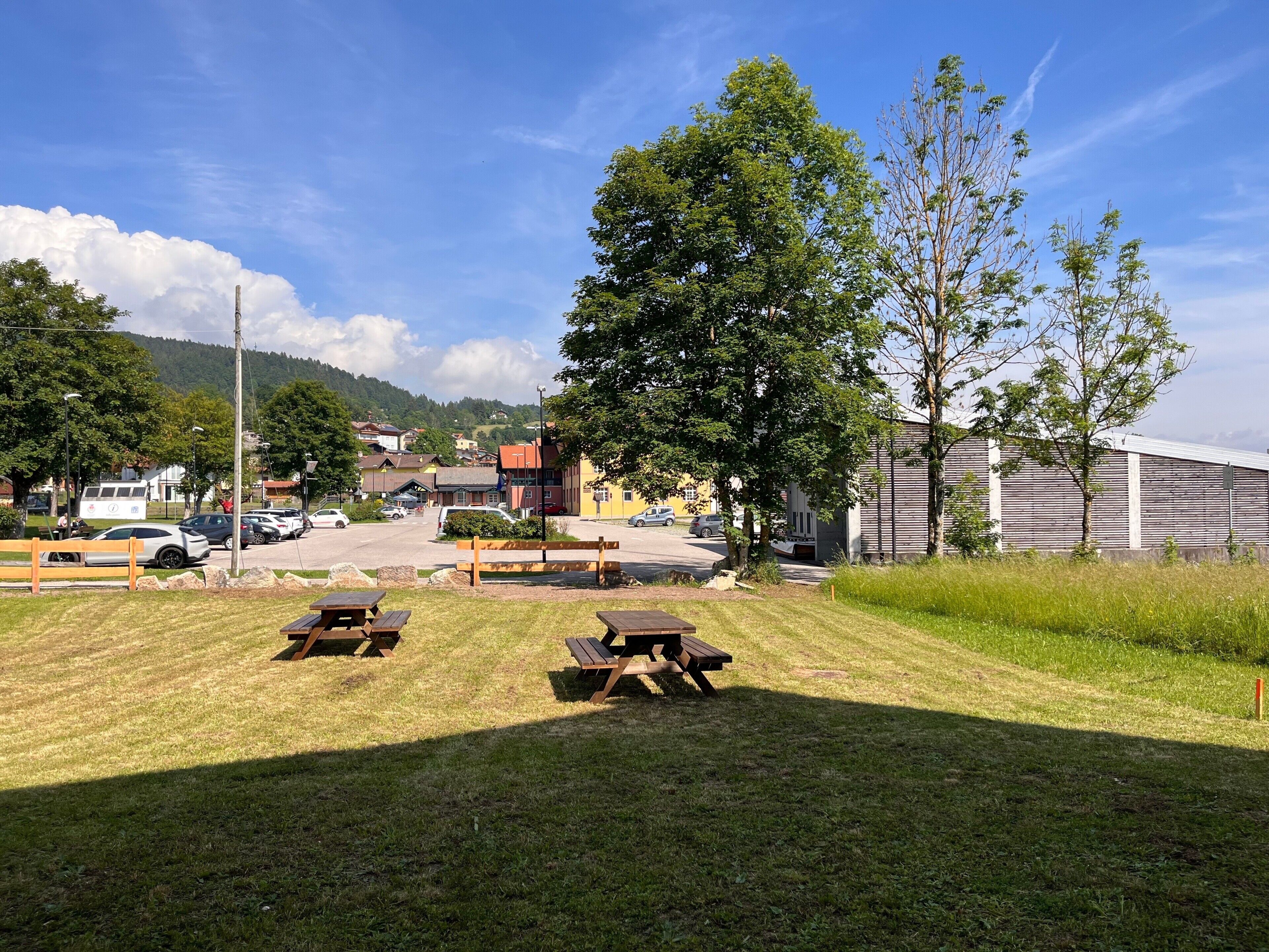 A picnic area with benches and picnic tables, a parking lot, and buildings in the background.