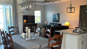 Un coin repas avec une table dressée pour six personnes, des chaises en bois, un ventilateur de plafond et un espace cuisine comprenant un évier et un plan de travail.