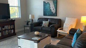 Un salon comprenant un grand téléviseur à écran plat, une table basse, un canapé et un fauteuil. Il y a une carte accrochée au mur et une lampe sur une table d’appoint.