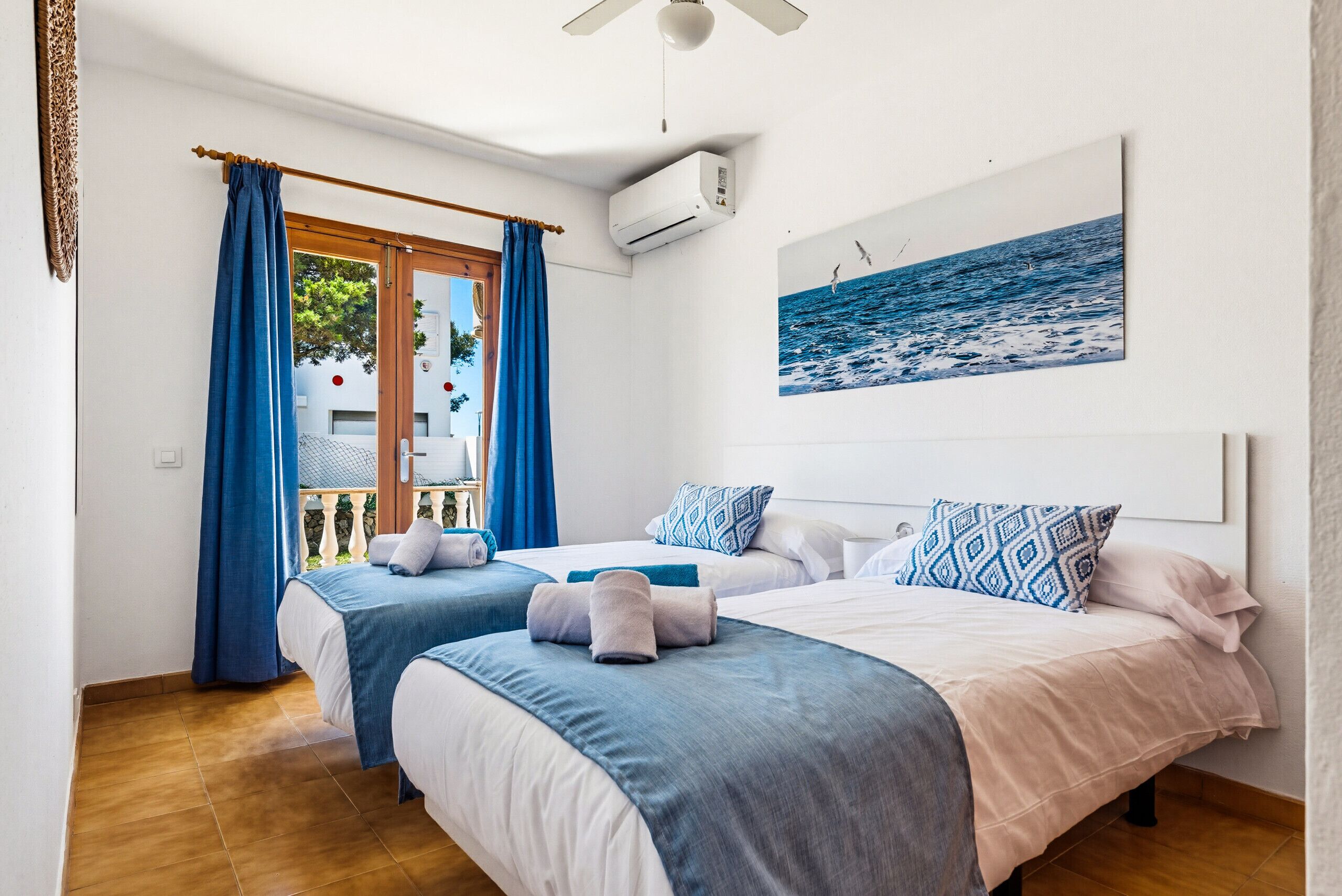 Habitación de hotel con dos camas, ventilador de techo, equipo de aire acondicionado, balcón con cortinas azules y un cuadro del mar.