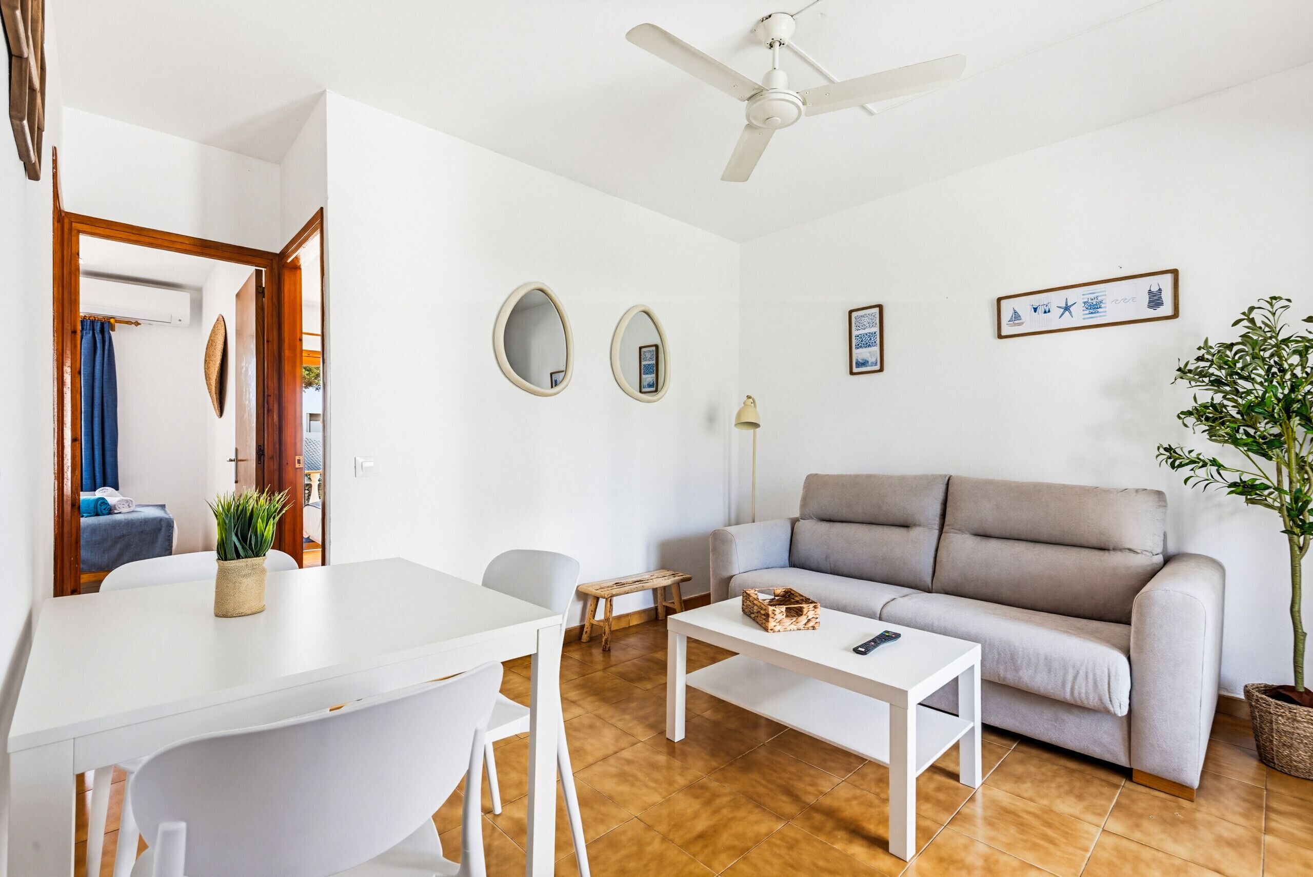A living room with a white sofa, a coffee table, a dining table with chairs, a ceiling fan, and a potted plant.
