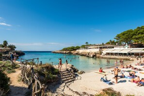 A beach with a wooden staircase leading down to the water, a sandy area with people sunbathing, and a rocky coastline.