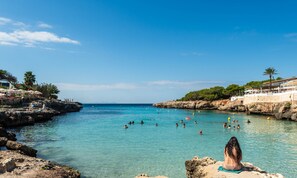 A coastal beach scene with clear blue waters, rocky shores, and a building with a terrace overlooking the sea.