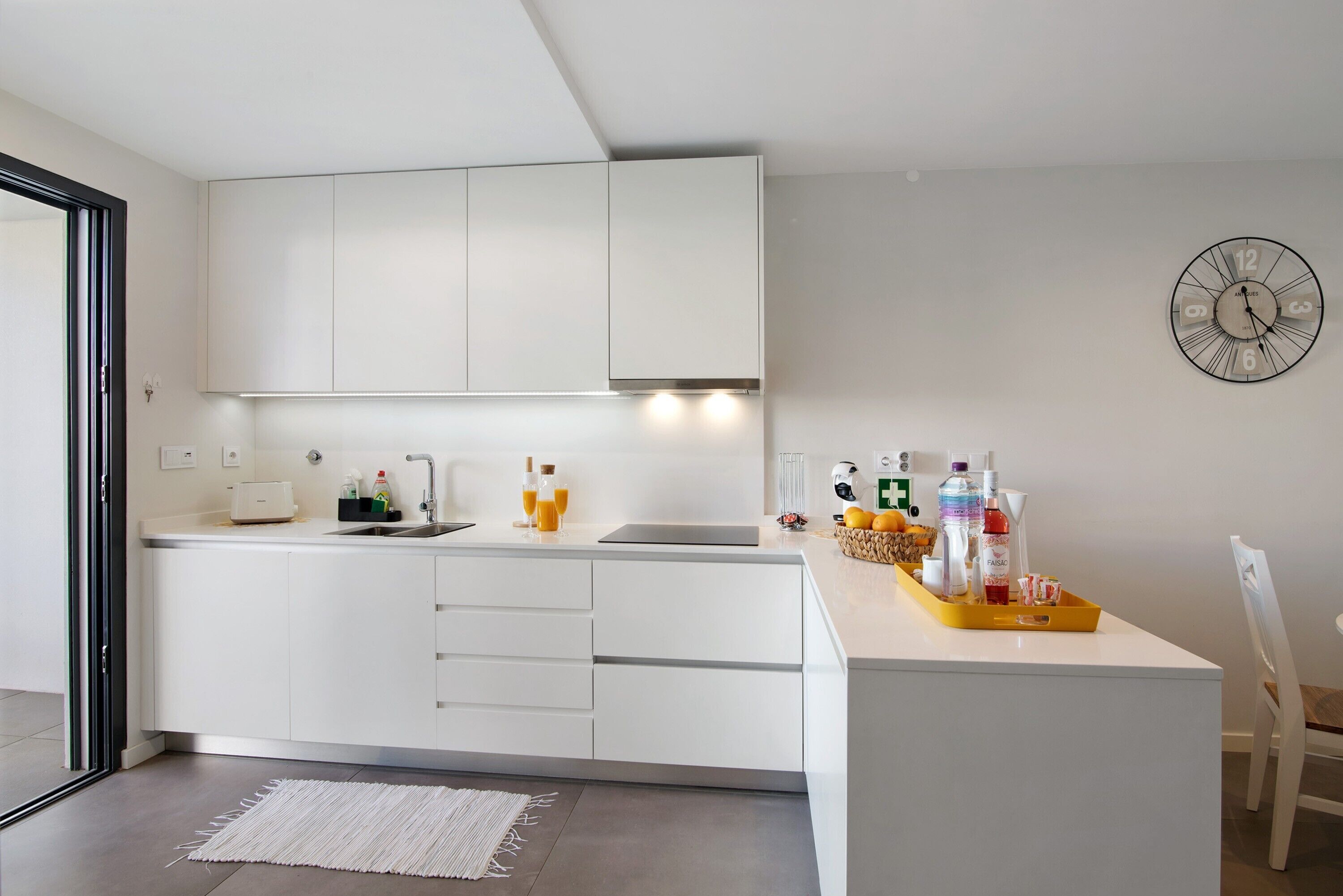 Een moderne keuken met witte kastjes, een inbouwoven, een spoelbak en een klok aan de muur.