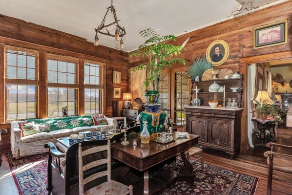 A traditional room with wooden walls, a sofa, a dining table with chairs, a chandelier, and a large plant.