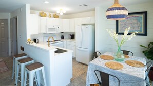 A modern kitchen with white cabinetry, a central island with stools, a dining table with woven placemats, and a hanging light fixture. - AK Getaway Too ,where the waves kiss the shore and worries melt away. (Port Aransas)