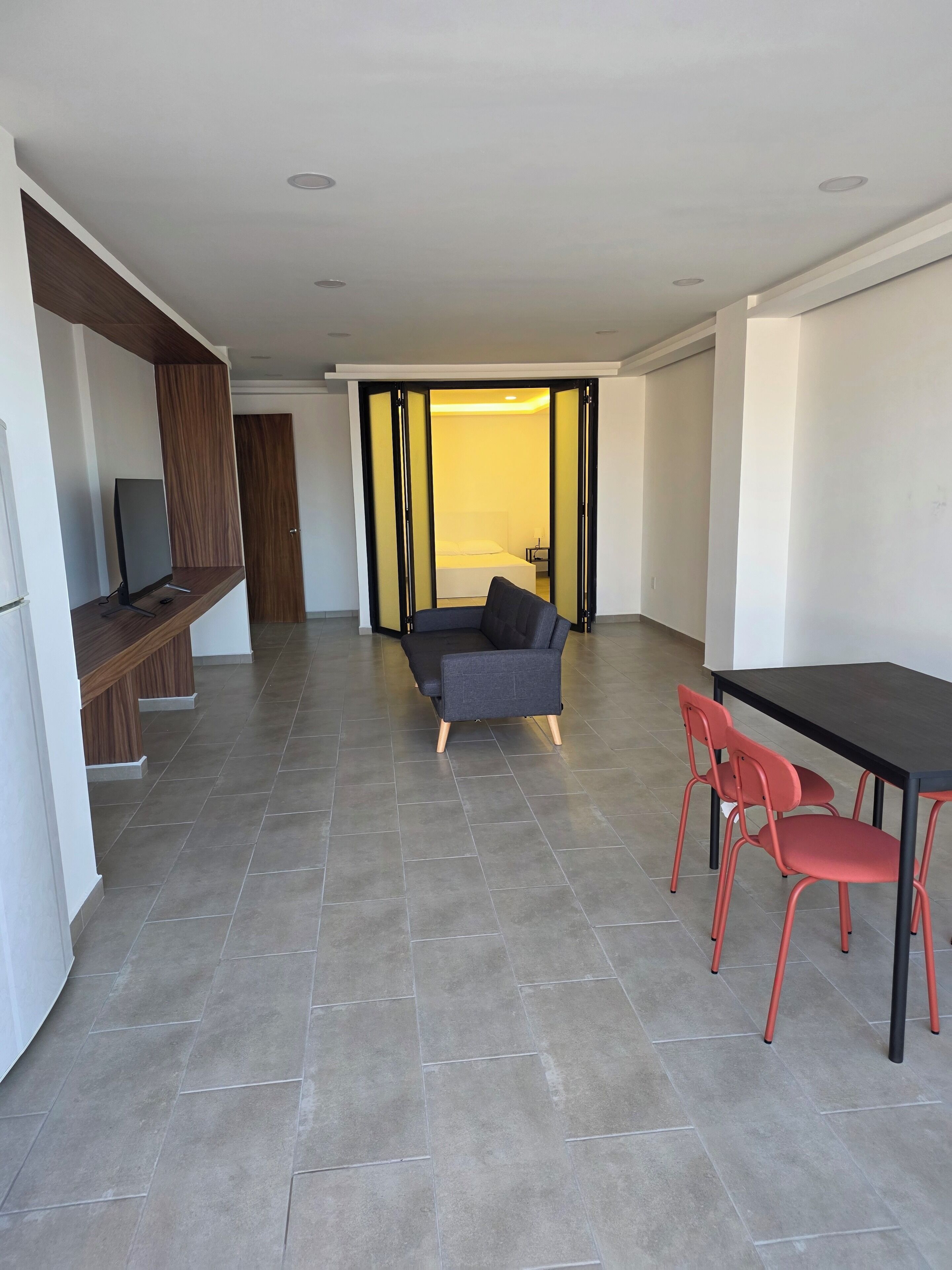 A modern living room with a sofa, a dining table with chairs, a TV on a wooden shelf, and a sliding door leading to a bedroom.