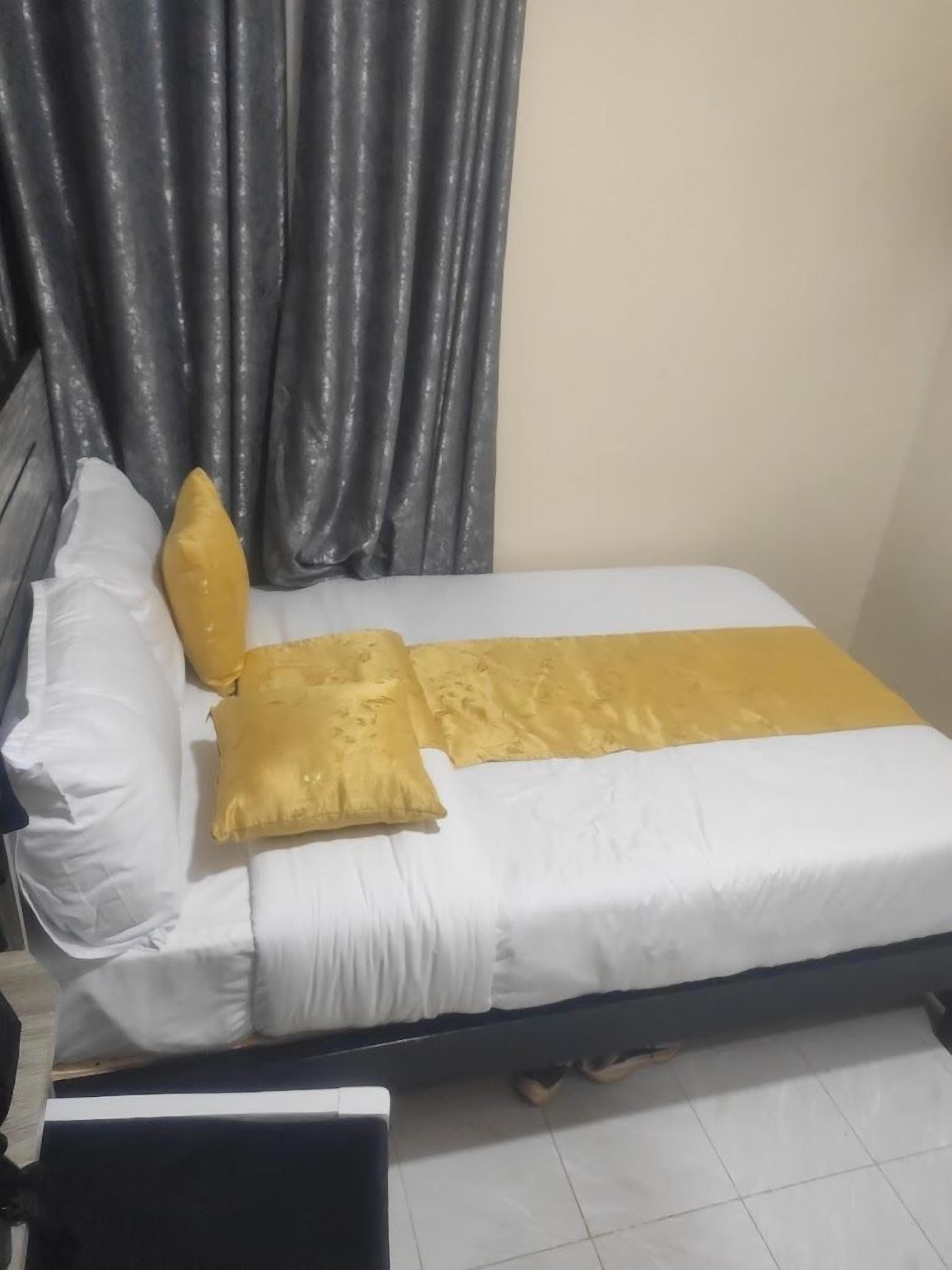 Una cama con ropa de cama blanca y dorada, cortinas grises y suelo de madera.