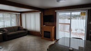 A living room with a sofa, a fireplace, a TV, and a view of a snowy landscape through a sliding door. - Chalet R&S on Lake Remy & Spa (Amherst)