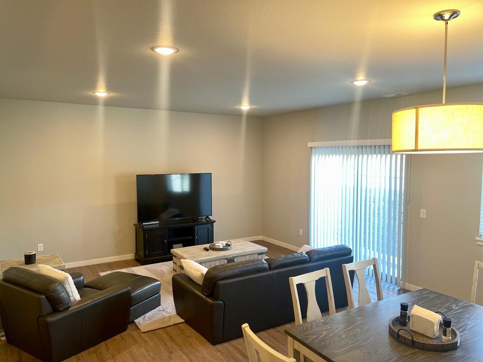 Een moderne woonkamer met een flatscreen televisie, donkere banken, een salontafel en een eethoek met stoelen en een houten tafel.