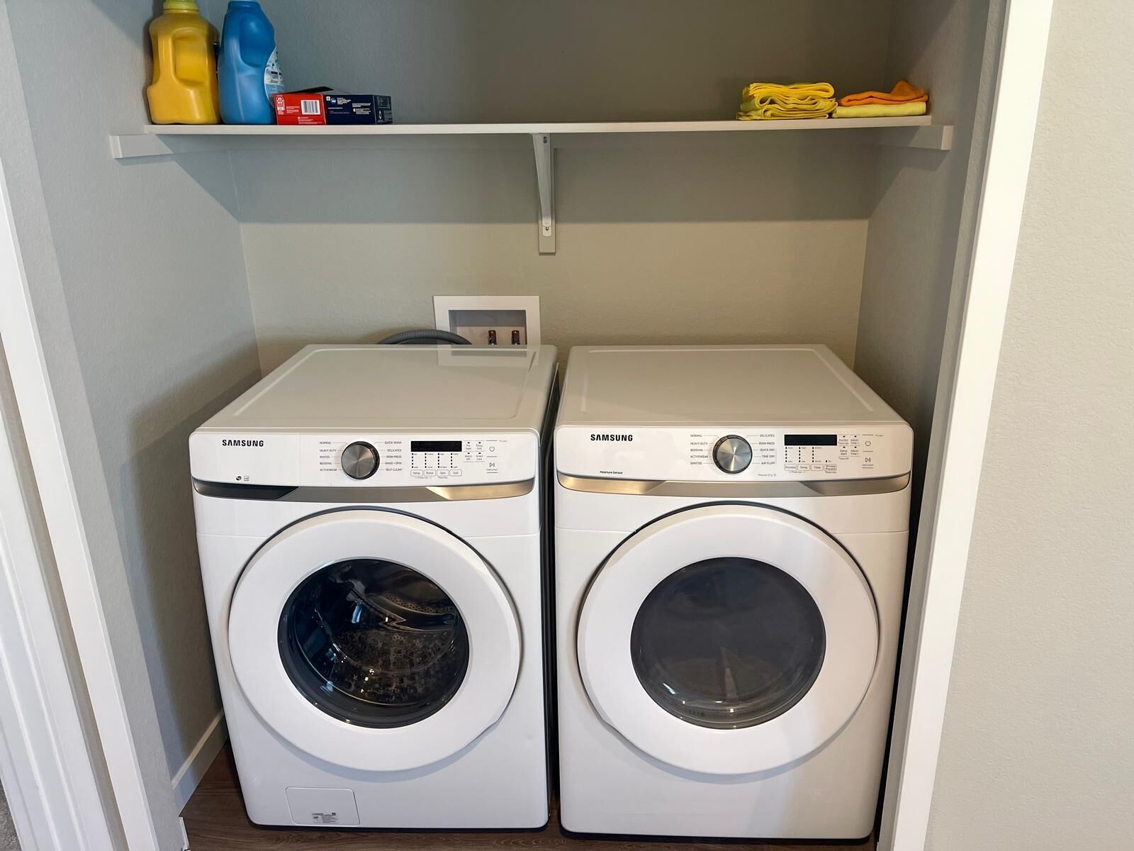 Two Samsung front-loading washing machines in a laundry room with a white shelf above containing cleaning supplies.