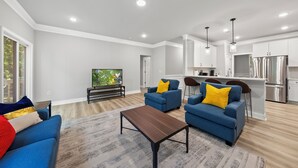 A modern living room with a sofa, a coffee table, and a TV. There is a kitchen area with a refrigerator and a dining table. - Woodland Retreat at Chapel Hill (Chapel Hill)