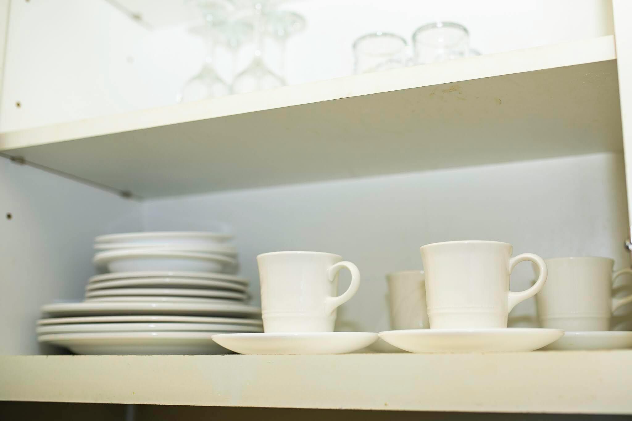A kitchen area with white cabinets and a shelf holding white cups and saucers.