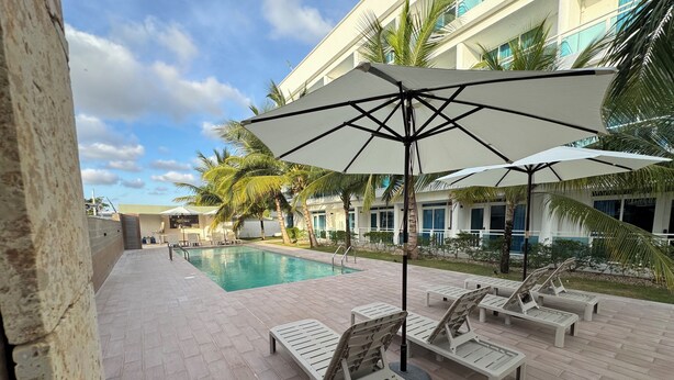 A poolside area with lounge chairs and umbrellas, a swimming pool, and a building with large windows and palm trees in the background. - Comfortable and modern studio (Punta Cana)