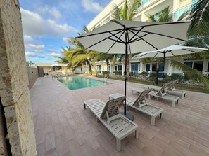 A poolside area with lounge chairs and umbrellas, a swimming pool, and a building with large windows and palm trees in the background. - Comfortable and modern studio (Punta Cana)