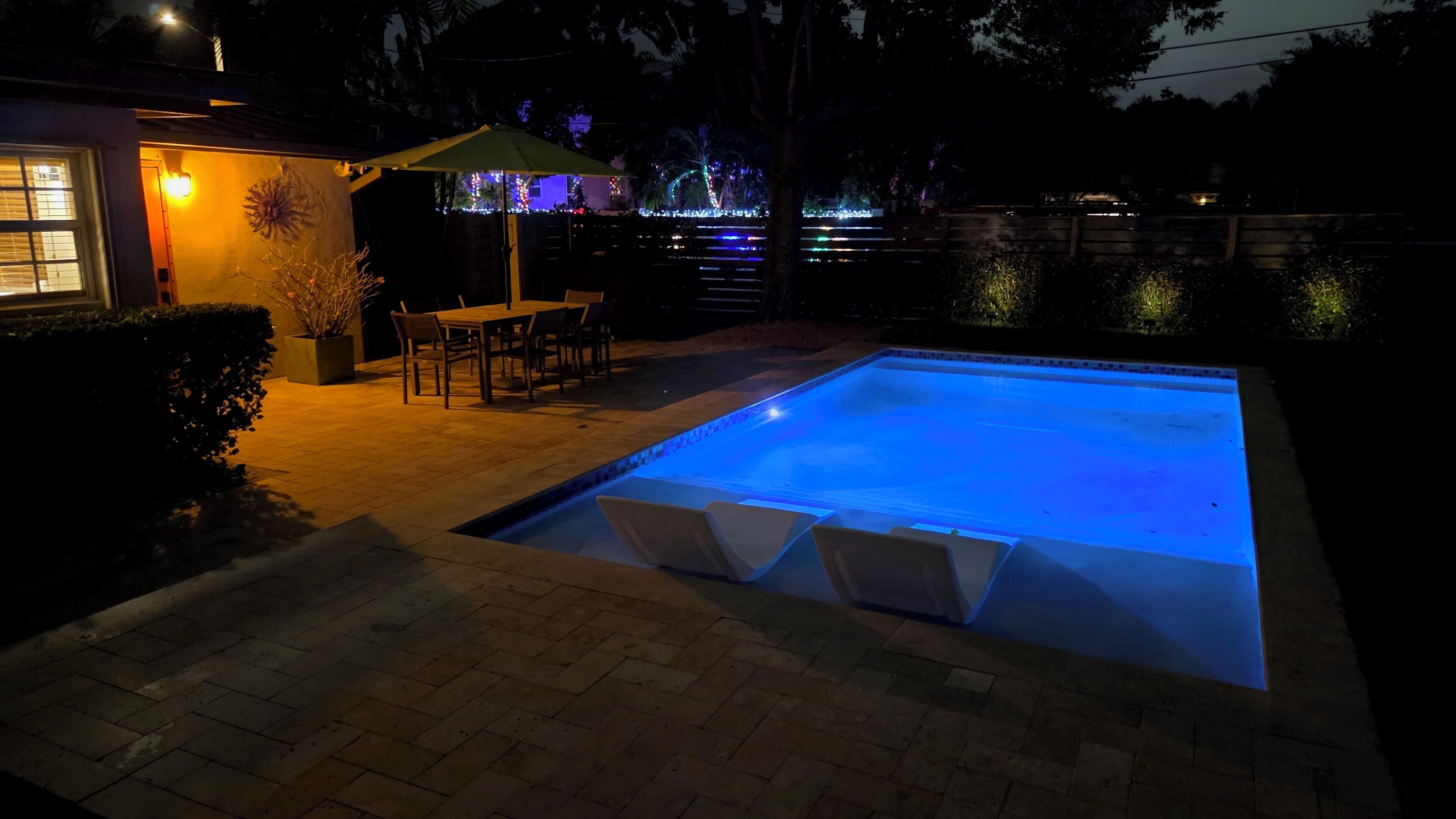 Une piscine éclairée de lumière bleue, une terrasse avec des chaises et une table.