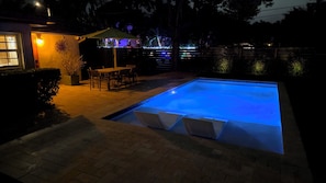 A swimming pool with a blue light, a patio with chairs, and a table.
