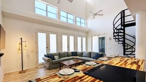 A spacious living room with a high ceiling, a large sofa set, a dining table with chairs, and a spiral staircase.