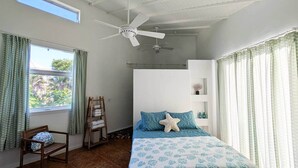 A bedroom with a bed, a ceiling fan, a window with curtains, a wooden chair, and a small shelf with decorative items.