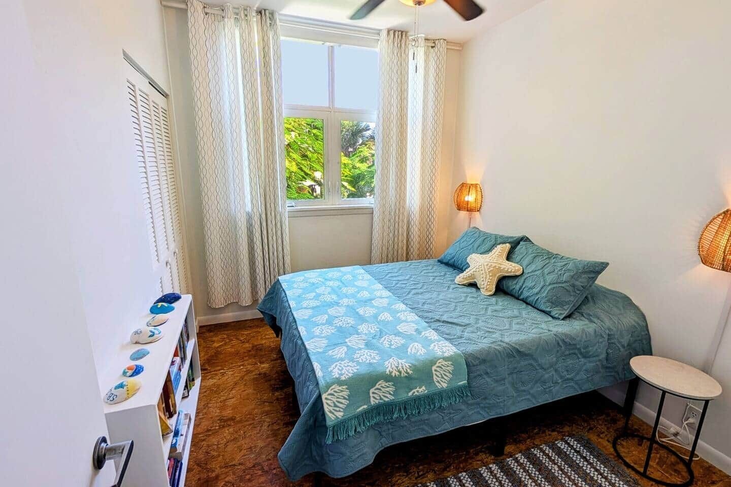 Een slaapkamer met een bed, gordijnen, een raam, een lamp en een klein tafeltje.
