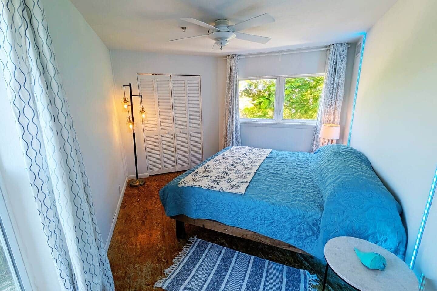 A bedroom with a bed, a ceiling fan, a window with curtains, a nightstand, and a floor lamp.