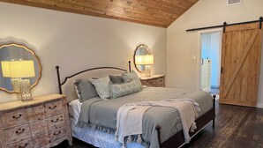 Un dormitorio con techo de madera, una cama con colcha gris, dos mesitas de noche con lámparas, un armario de madera y una puerta corredera estilo granero.