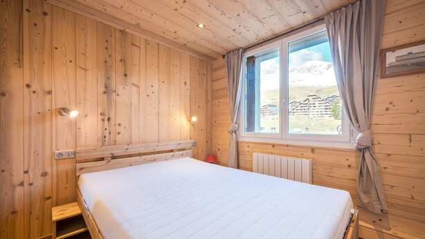 Ein Zimmer mit Holzboden, einem Bett, einem Fenster mit Vorhängen und Blick auf ein Gebäude.