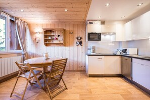 Une pièce aux murs lambrés, avec une table ronde en bois et des chaises, une cuisine dotée de meubles blancs et d’appareils électroménagers, et une fenêtre agrémentée de rideaux.