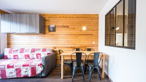 A room with a wooden wall, a dining table with chairs, a sofa with a patterned blanket, and a window. - Apartment 'Vacation Rental' with Mountain View and Balcony (Saint François Longchamp)