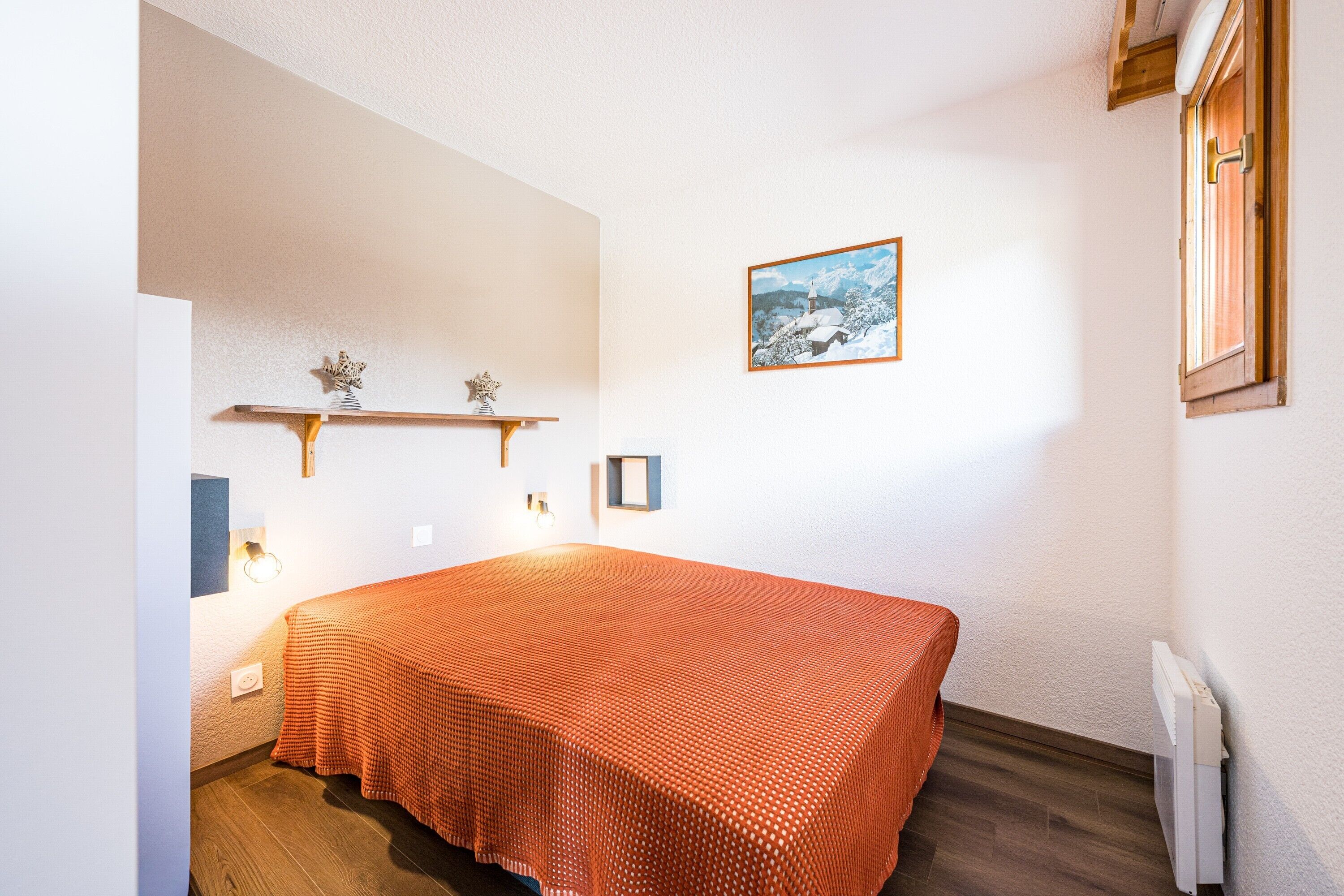 Ein Schlafzimmer mit Bett, einer an der Wand befestigten Ablage mit Dekoration, einem gerahmten Bild und einem Heizkörper.
