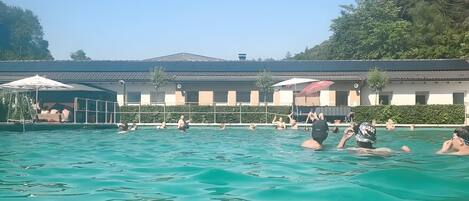 Une piscine avec des personnes, un bâtiment avec un toit en tuiles et de la verdure en arrière-plan.