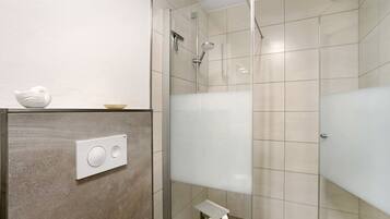 A modern bathroom with a glass shower enclosure, a toilet paper holder, and a wall-mounted control panel.