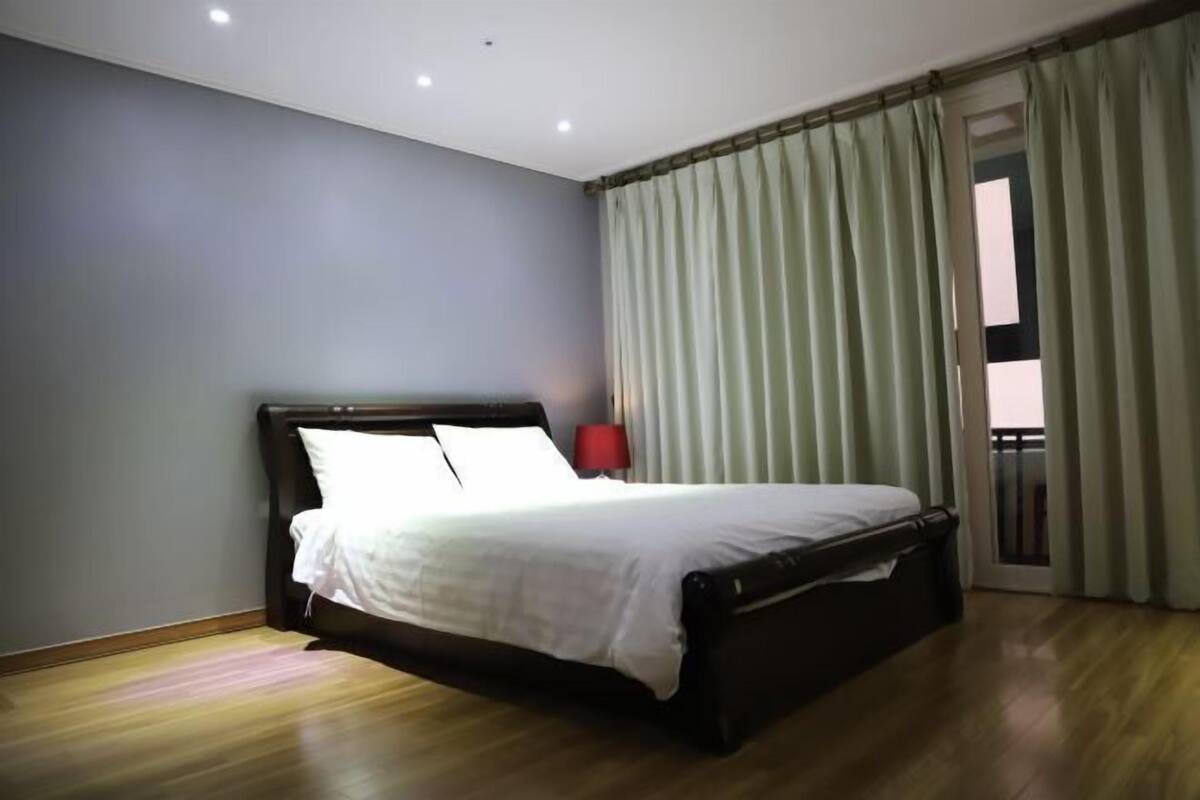 A bedroom with a bed, a nightstand, a red lamp, and a view of the outside through a window with curtains.