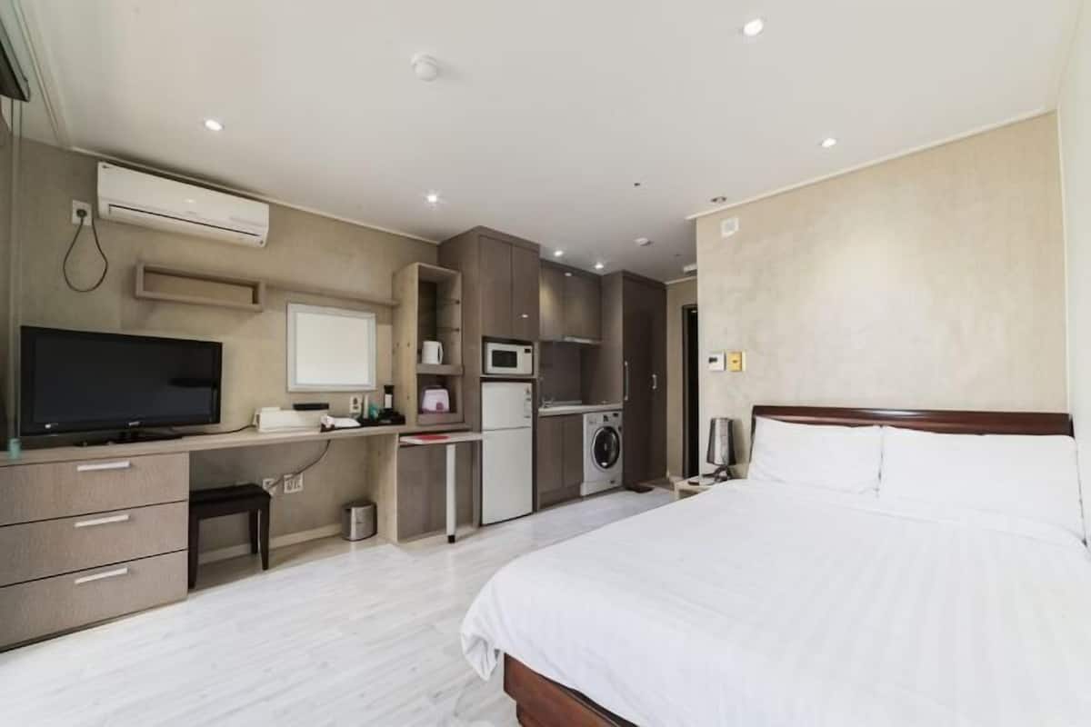 A hotel room with a bed, a desk with a computer, a TV, a microwave, and a small kitchenette.