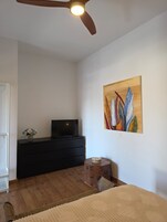 Una habitación con ventilador de techo, un televisor de pantalla plana sobre un tocador negro, un cuadro en la pared y una alfombra tejida en el suelo.