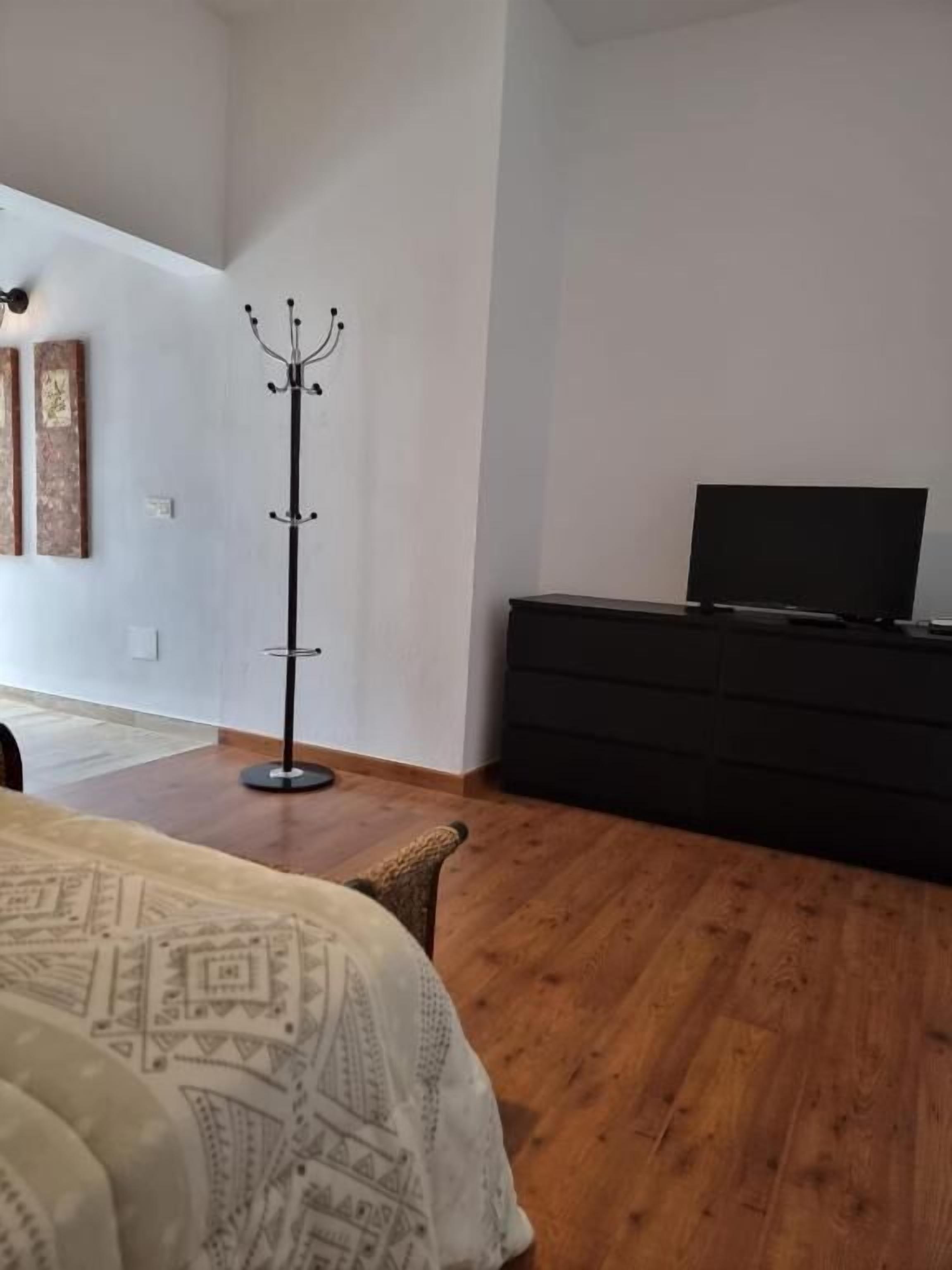 Un dormitorio con una cama, perchero, televisor sobre un tocador y piso de madera.