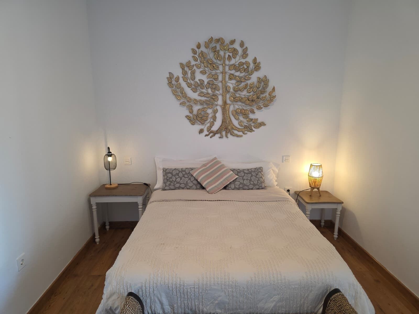 Une chambre avec un grand lit, deux tables de chevet avec des lampes, une décoration murale représentant un arbre et un plancher en bois.