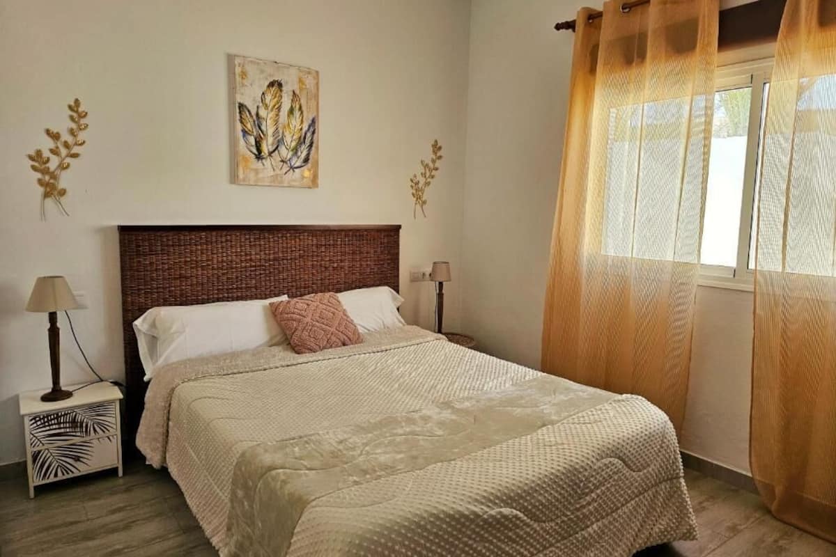 Un lit bien fait, avec une couvre-lit à motifs, une tête de lit en bois et une table de chevet surmontée d’une lampe.
