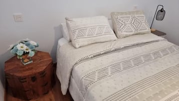 Un dormitorio con una cama, dos almohadas, una mesita de noche con una lámpara y un taburete de madera con un arreglo floral.