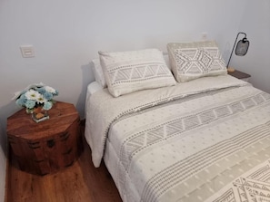 A bedroom with a bed, two pillows, a bedside table with a lamp, and a small wooden stool with a flower arrangement.