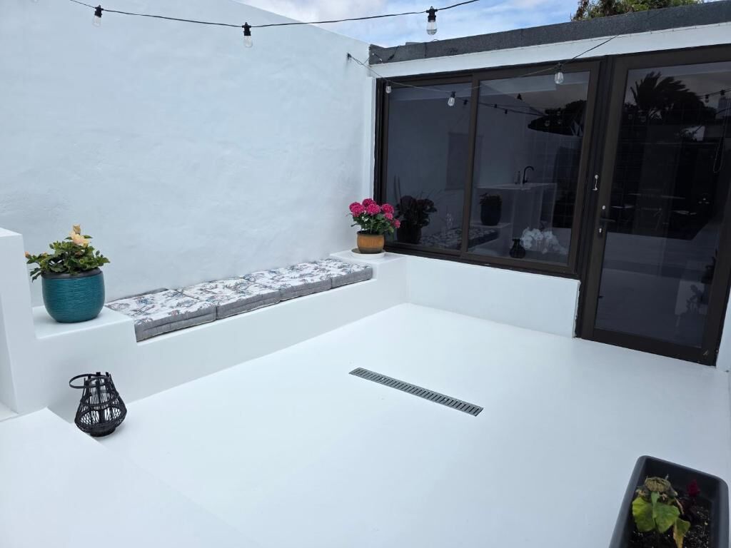 A rooftop terrace with a white floor, a potted plant, a small black lamp, and a window with a view of the interior.