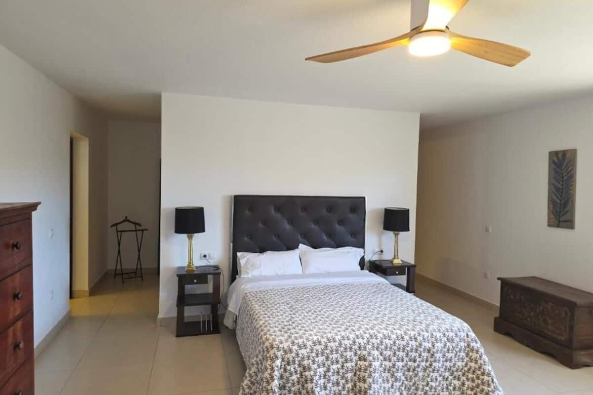 Une chambre à coucher comprenant un lit, deux tables de chevet, un ventilateur de plafond, une commode et un portant à vêtements.